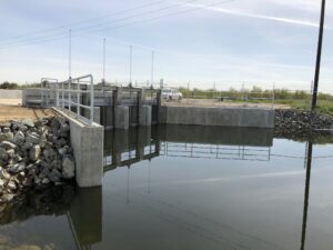 photo of concrete turnout structure, water, rock-lined channel, and farmland in the background.